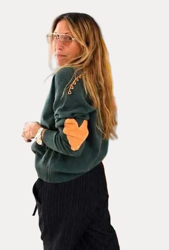 A woman with long light brown hair and glasses stands in front of a colorful abstract painting. She is wearing a dark green sweater with orange accents and black pants.