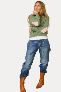 A woman stands in a room wearing a sage green knit sweater with pink elbow patches, blue jeans with paint splatters, and brown leather boots. She is posed in front of a white wall with colorful abstract paint strokes and a light wood floor.