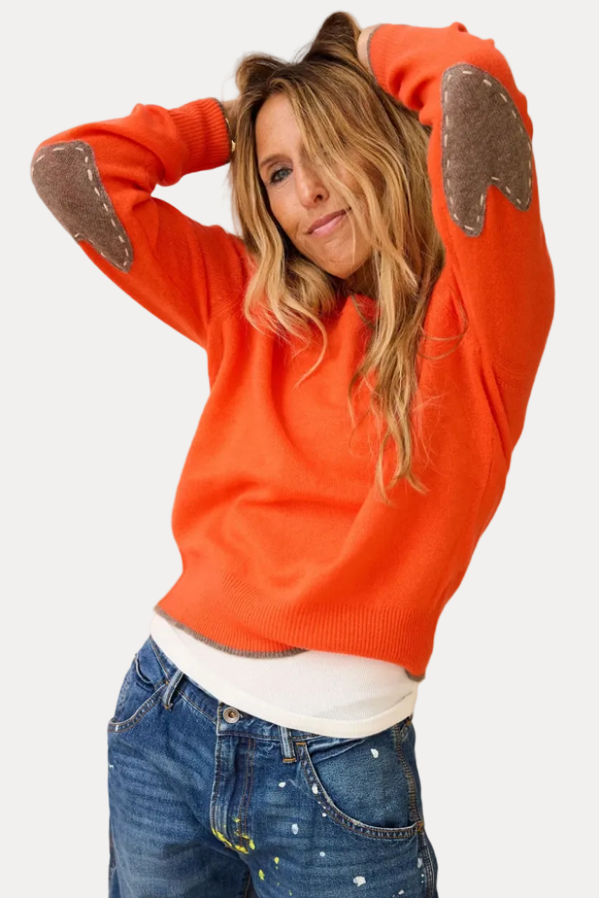 A woman wearing a bright orange knit sweater with brown heart-shaped patches on the shoulders and elbows, styled with blue jeans and a white undershirt.