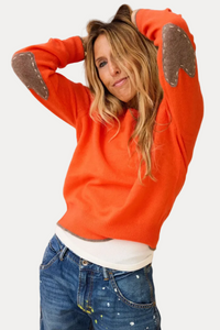 A woman wearing a bright orange knit sweater with brown heart-shaped patches on the shoulders and elbows, styled with blue jeans and a white undershirt.