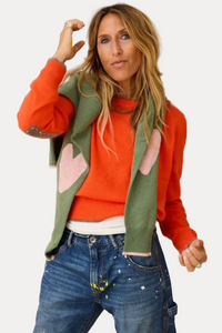 A woman wearing a vibrant orange crewneck sweater layered under a green knit cardigan with pink heart patches, paired with blue jeans, posing in front of a modern abstract wall.