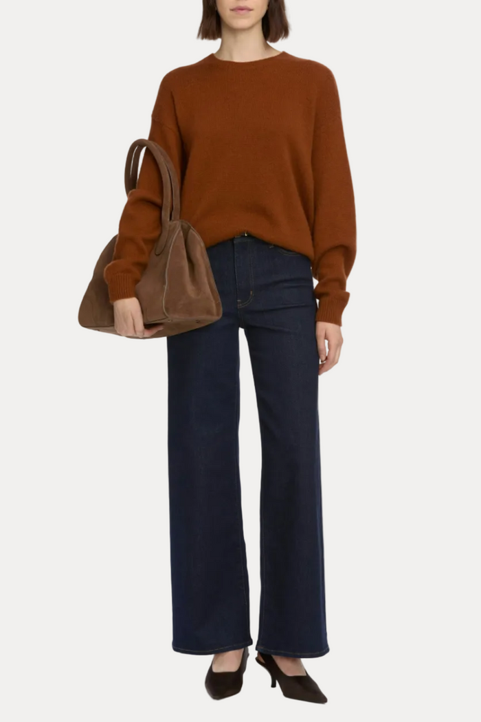 Person wearing a brown sweater and dark blue jeans, holding a brown handbag on a white background