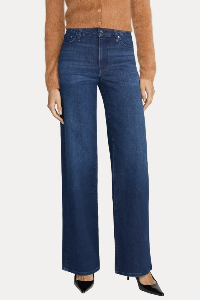 A model wearing dark blue wide-leg denim jeans, styled with a brown cardigan and black pointed-toe heels, photographed against a neutral light gray studio background.