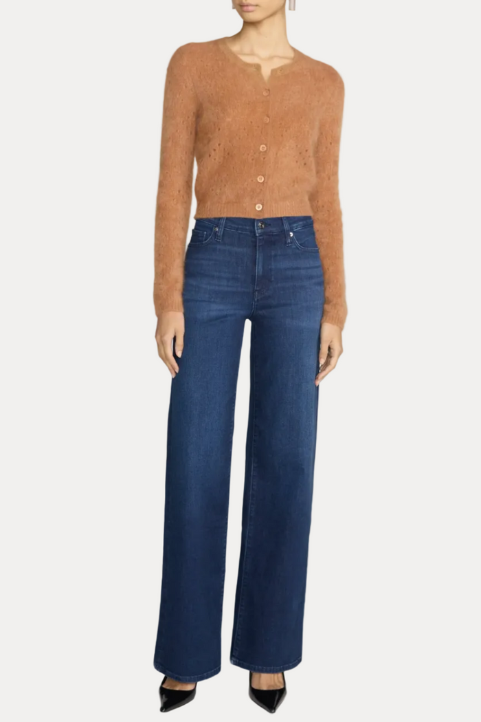 A model wearing a cropped, fuzzy tan button-up cardigan and high-waisted, wide-leg blue jeans, paired with black pointed-toe heels, against a light gray studio background.