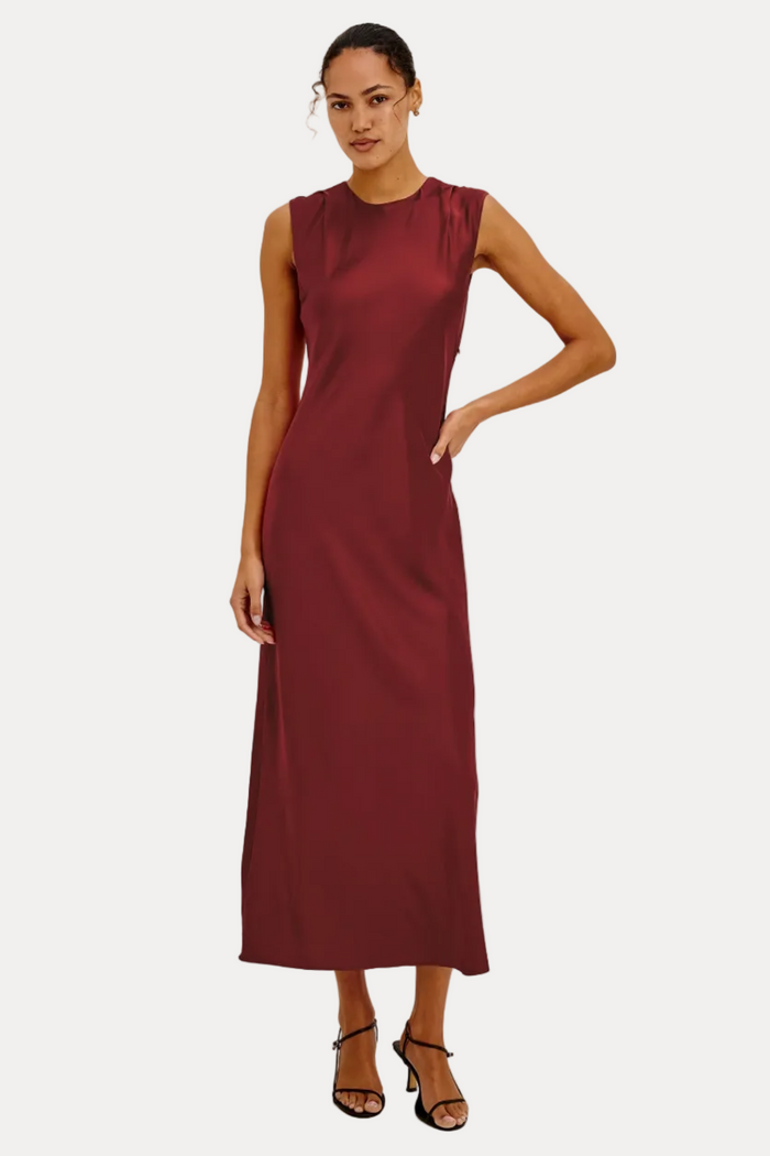 A woman modeling a sleeveless, high-neck, midi-length satin dress in a deep burgundy red color, paired with black strappy heels, against a plain white studio background.