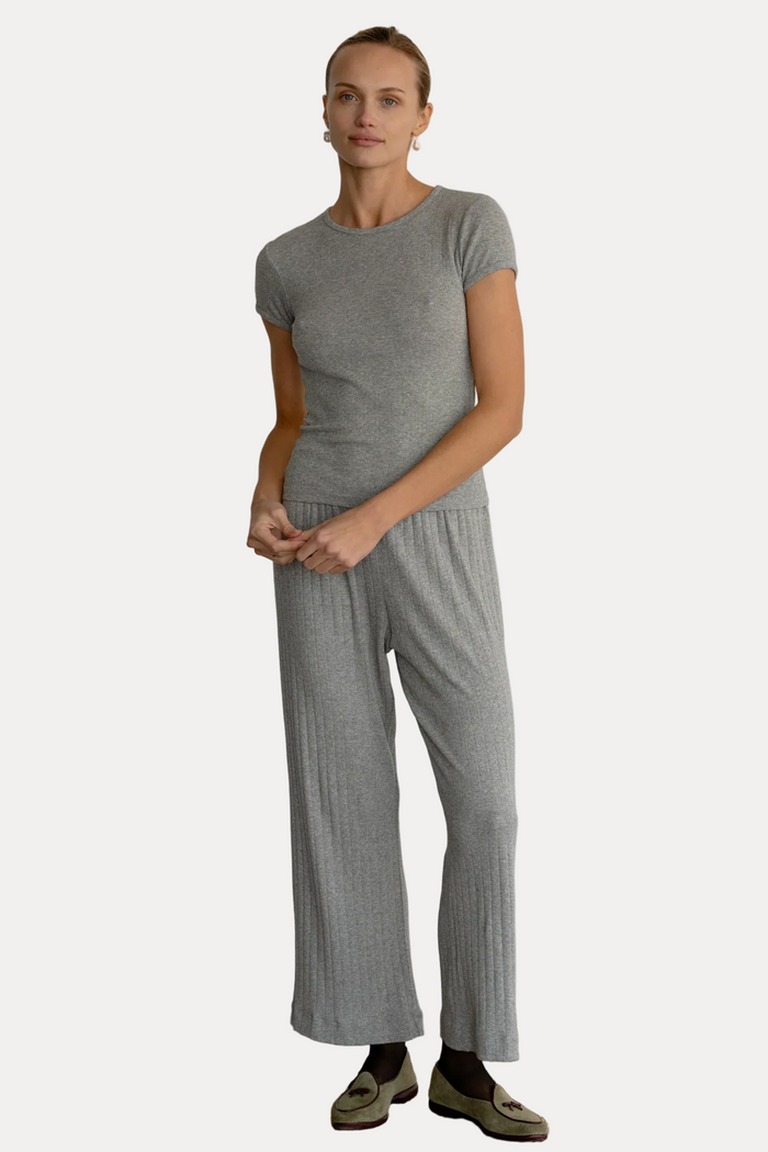 A woman modeling a light gray short-sleeve crewneck top and matching wide-leg pleated pants, standing against a plain white wall on a textured beige floor.