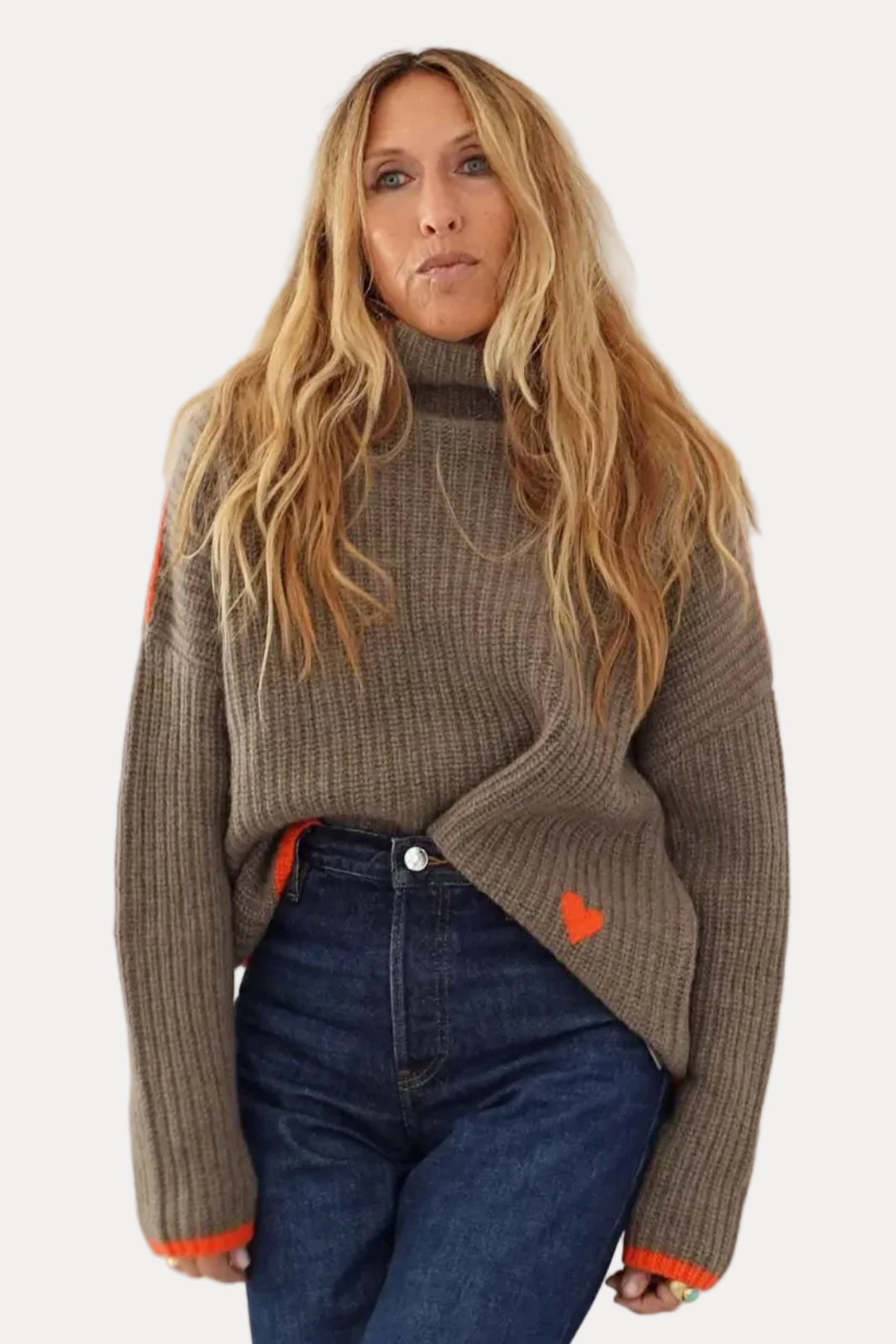 A woman with long blonde hair wearing a brown ribbed knit turtleneck sweater with bright orange accents and dark blue jeans, standing against a plain off-white wall.