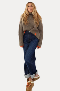 A woman stands against a plain white wall wearing a ribbed taupe-brown turtleneck sweater with bright orange cuffs, dark blue jeans, and tan slip-on shoes with orange laces.