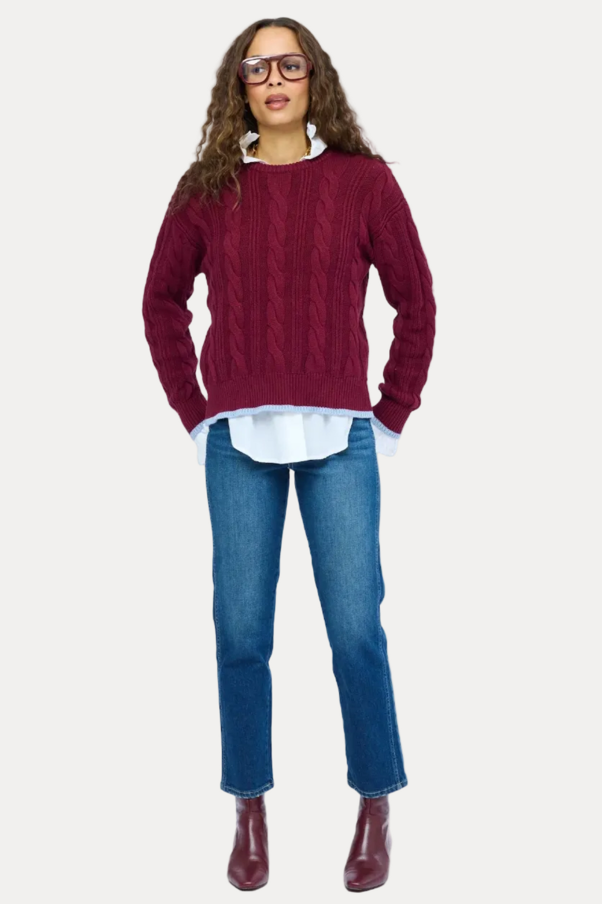 A woman modeling a burgundy cable-knit sweater over a white collared shirt, paired with blue jeans and burgundy ankle boots, against a plain white background.