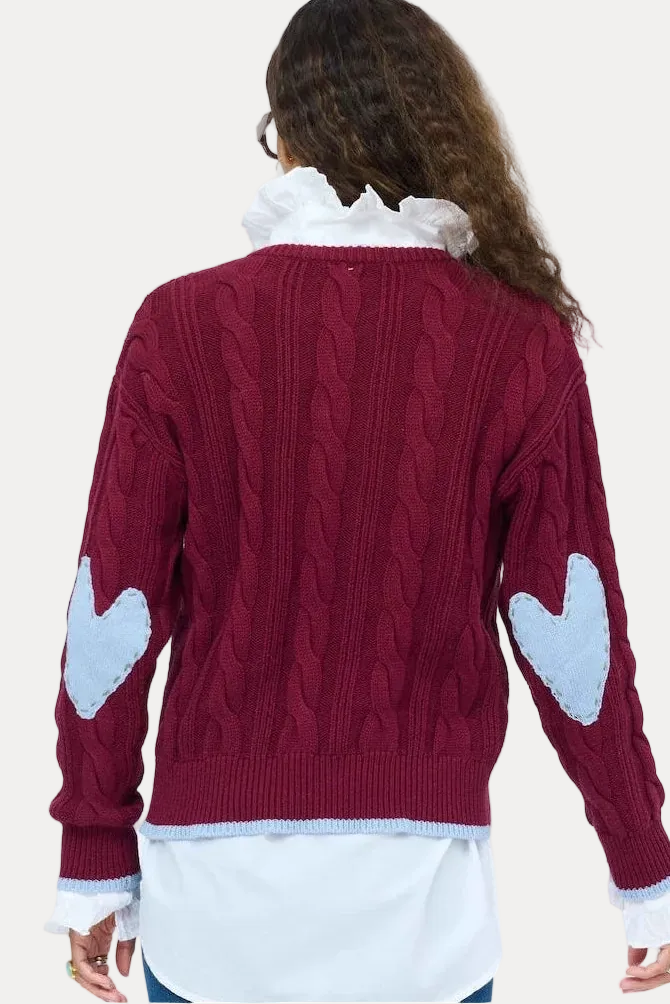 A woman viewed from behind wearing a burgundy cable-knit sweater with light blue heart patches on the sleeves, layered over a white collared shirt and blue jeans, against a plain white background.