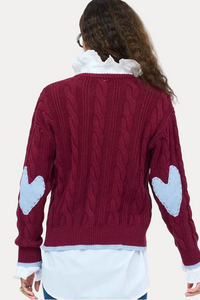 A woman viewed from behind wearing a burgundy cable-knit sweater with light blue heart patches on the sleeves, layered over a white collared shirt and blue jeans, against a plain white background.