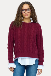 A woman modeling a burgundy cable-knit sweater over a white collared shirt, paired with blue jeans and burgundy ankle boots, against a plain white background.