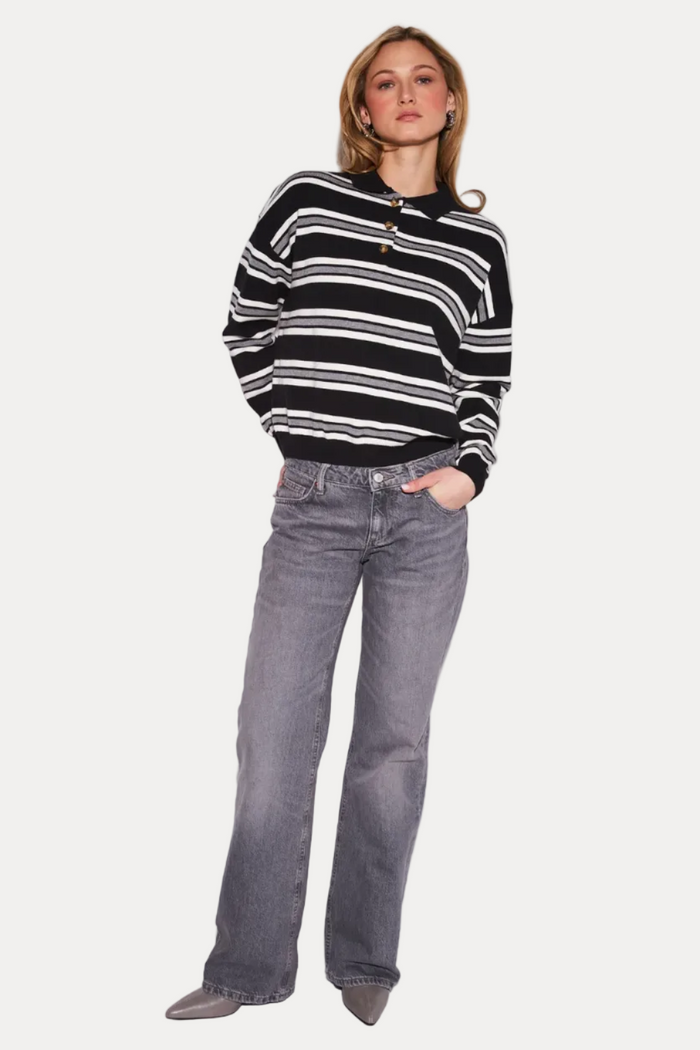 A woman modeling a black and white horizontally striped knit polo sweater with gray wide-leg jeans and gray pointed shoes, standing against a plain light gray studio background.