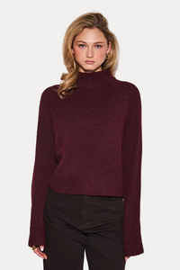 A woman modeling a dark burgundy knit mock neck sweater with long sleeves, paired with black pants, against a plain light gray background.