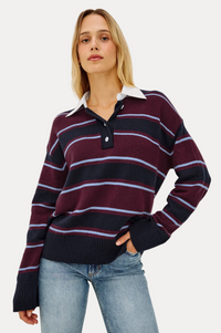 A woman modeling a long-sleeve rugby-style knit sweater with horizontal stripes in burgundy, navy, and light blue, featuring a white collar and a dark navy button placket, paired with light-wash blue jeans against a plain light gray background.