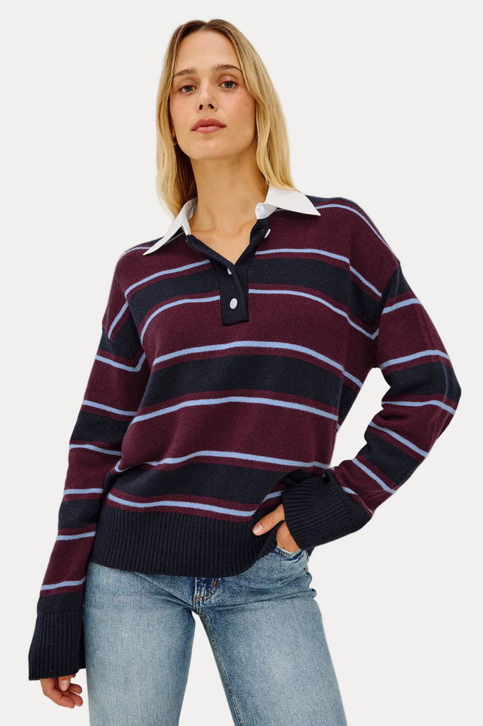 A woman modeling a long-sleeve rugby-style knit sweater with horizontal stripes in burgundy, navy, and light blue, featuring a white collar and a dark navy button placket, paired with light-wash blue jeans against a plain light gray background.
