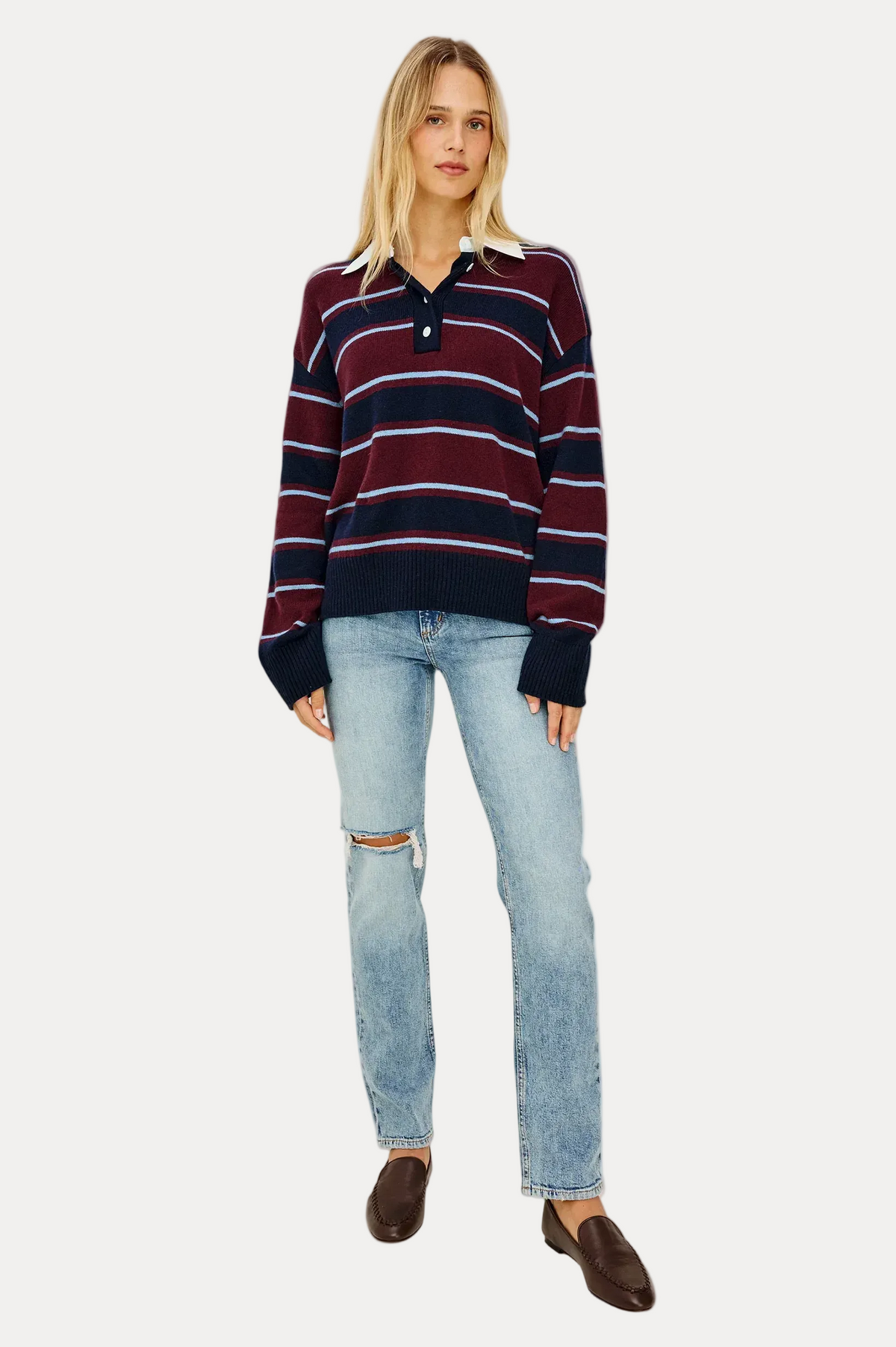 A woman modeling a long-sleeve knit polo sweater with horizontal burgundy, navy, and white stripes, paired with light-wash distressed jeans and dark brown loafers, against a plain off-white studio background.
