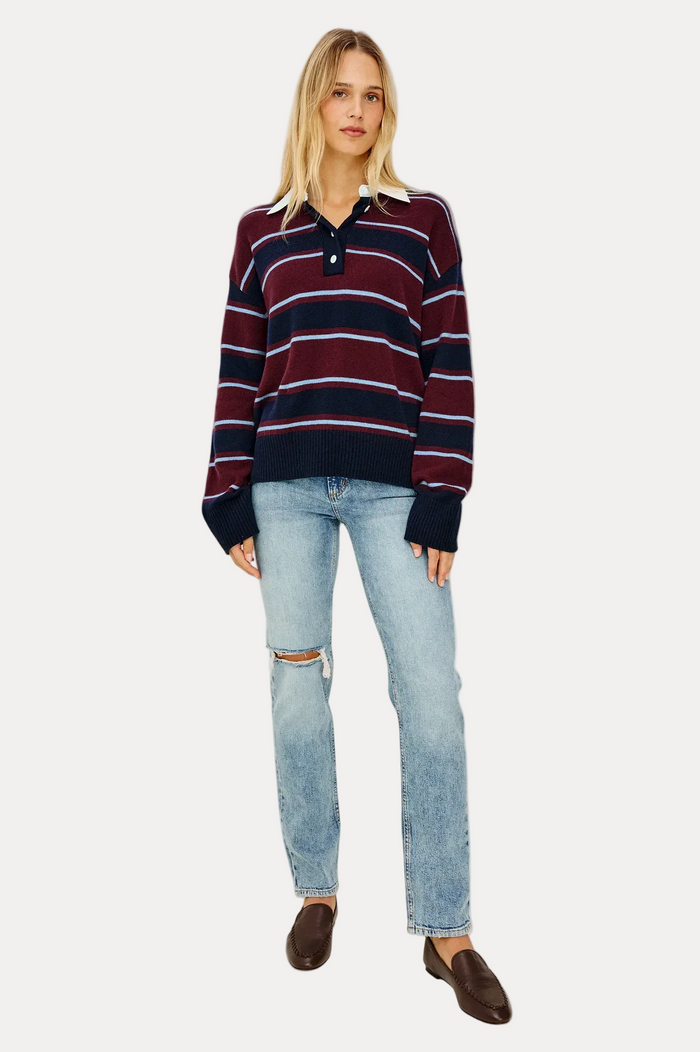 A woman modeling a long-sleeve knit polo sweater with horizontal burgundy, navy, and white stripes, paired with light-wash distressed jeans and dark brown loafers, against a plain off-white studio background.