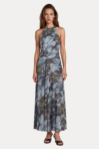 A woman modeling a sleeveless halter-neck maxi dress with a gray, blue, and brown abstract tie-dye pattern, standing against a light gray studio background.