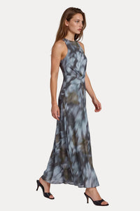 A woman modeling a sleeveless, high-neck, floor-length maxi dress with an abstract gray, blue, and brown watercolor-like print, paired with black heeled sandals, against a light gray studio background.