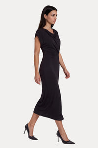 A woman modeling a black midi dress with a draped cowl neckline and cap sleeves, paired with black pointed-toe heels, against a light gray studio background.