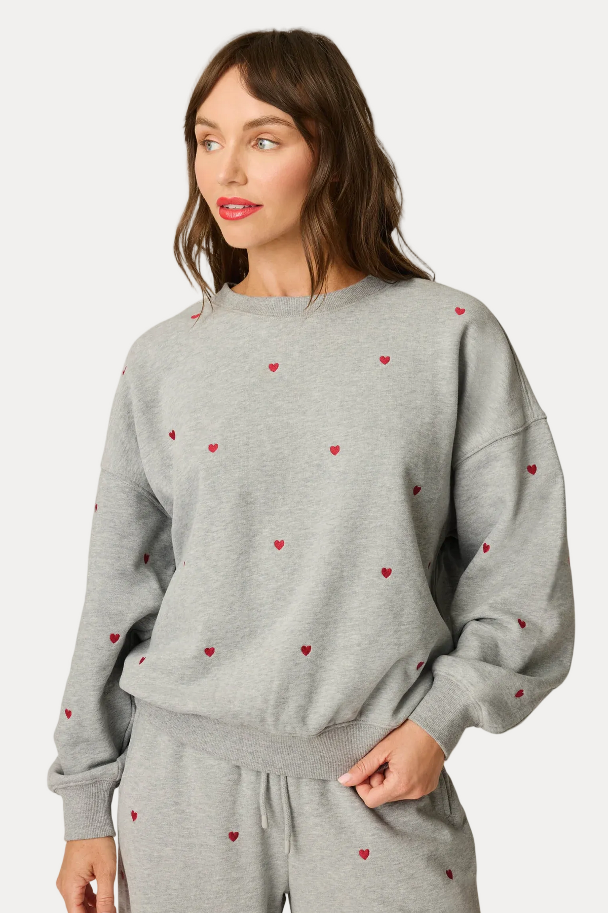 A woman modeling a light gray sweatshirt and matching sweatpants set, both decorated with small red embroidered hearts, against a plain off-white studio background.