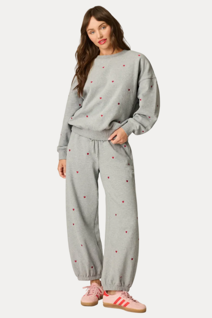 A woman modeling a light gray two-piece loungewear set with small red heart patterns, consisting of a crewneck sweatshirt and matching jogger pants, paired with pink sneakers against a light gray studio background.
