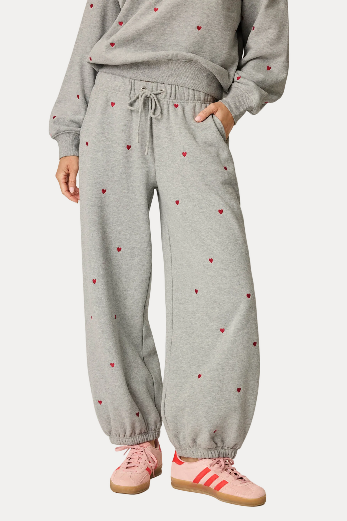 A person wearing gray jogger sweatpants with small red heart prints, paired with a matching gray sweatshirt and pink sneakers, shown from the chest down against a plain light background.