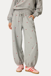 A person wearing gray jogger sweatpants with small red heart prints, paired with a matching gray sweatshirt and pink sneakers, shown from the chest down against a plain light background.