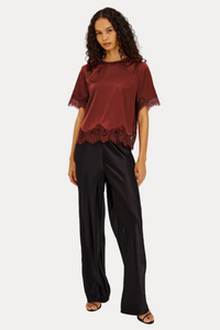 A woman modeling a short-sleeve maroon satin top with lace trim and black wide-leg satin pants against a light gray studio background.