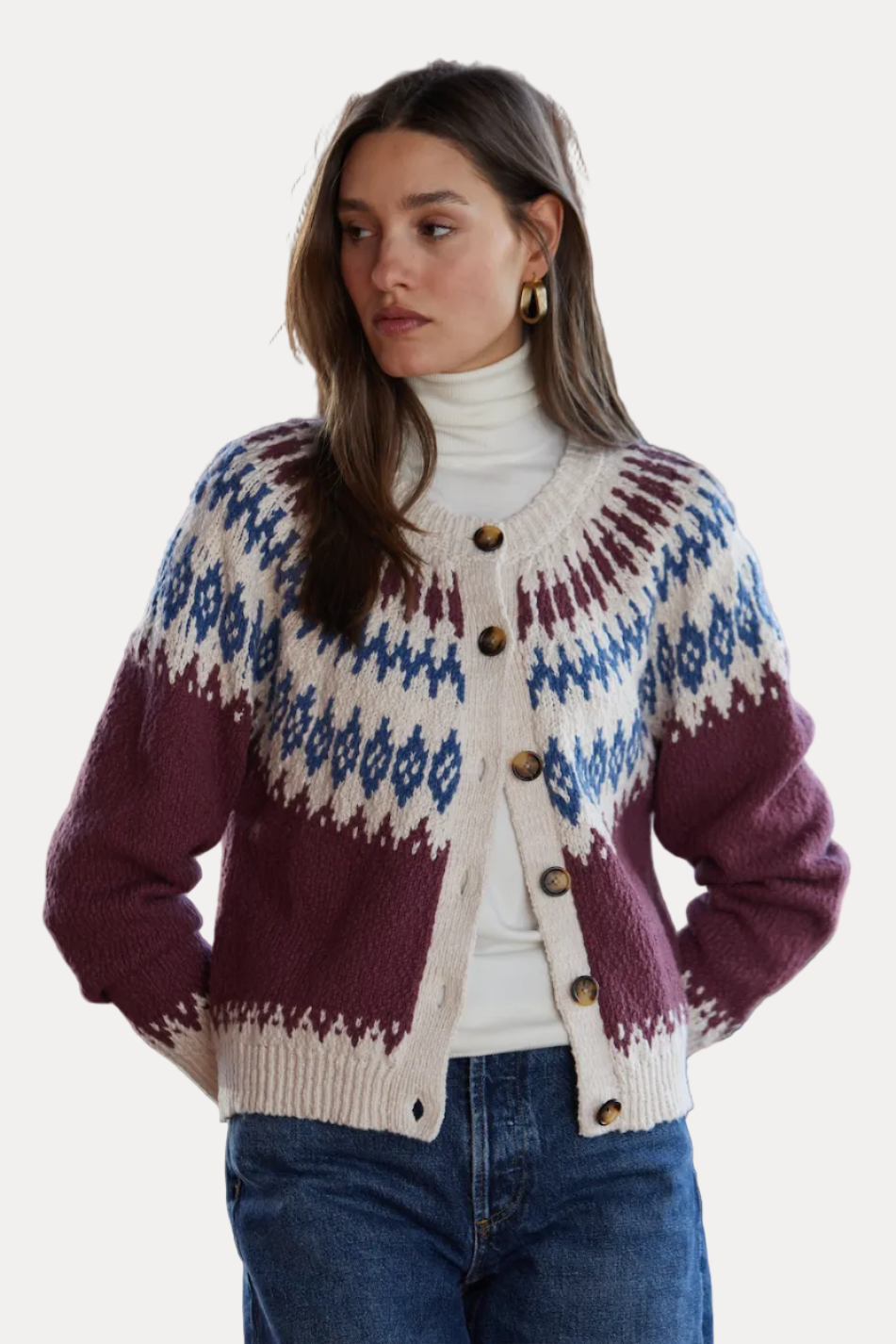 A woman wearing a button-front knit cardigan with a cream base, blue geometric patterns, and deep burgundy lower panels, styled over a white turtleneck and blue jeans in front of a brick wall.