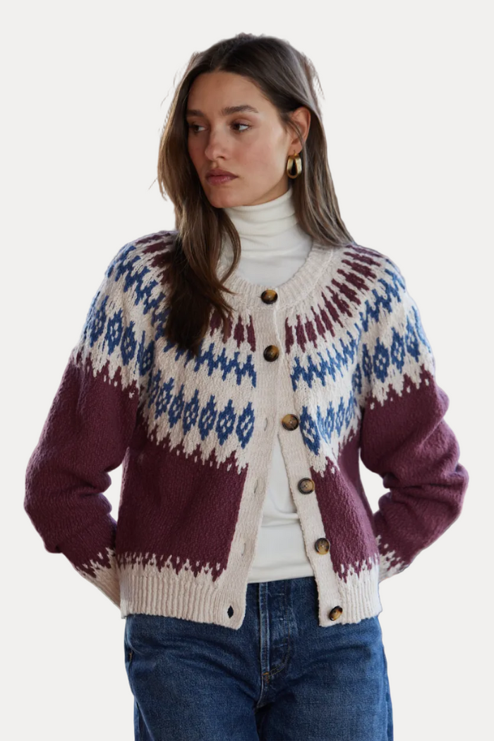 A woman wearing a button-front knit cardigan with a cream base, blue geometric patterns, and deep burgundy lower panels, styled over a white turtleneck and blue jeans in front of a brick wall.