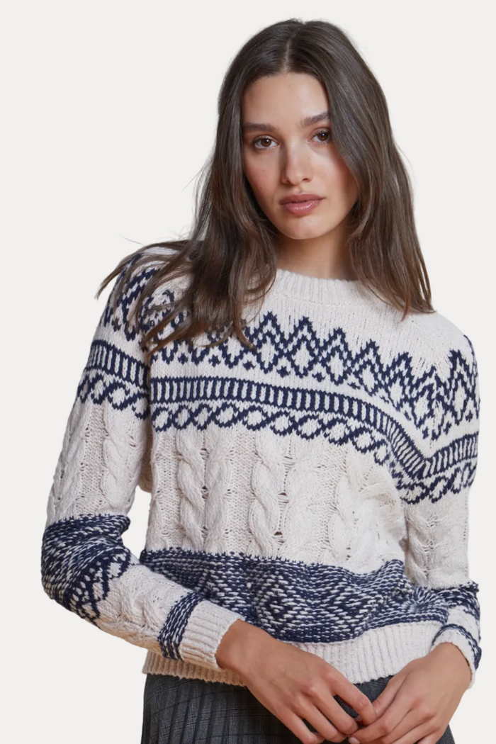 A woman wearing a cream and navy blue cable-knit sweater with a Fair Isle pattern, posed against a neutral light gray background.