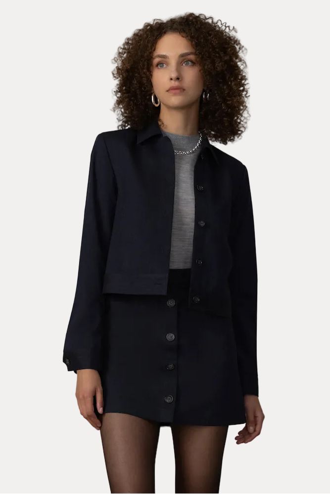 A model stands in front of a beige curtain backdrop wearing a dark navy cropped jacket and matching button-front mini skirt over a gray mock-neck top, paired with gray socks and black loafers.