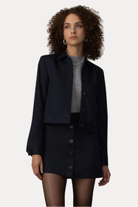A model stands in front of a beige curtain backdrop wearing a dark navy cropped jacket and matching button-front mini skirt over a gray mock-neck top, paired with gray socks and black loafers.