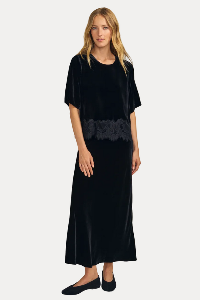 A woman modeling a black velvet midi dress with short sleeves and a black lace waist detail, paired with black flats, against a solid beige studio background.
