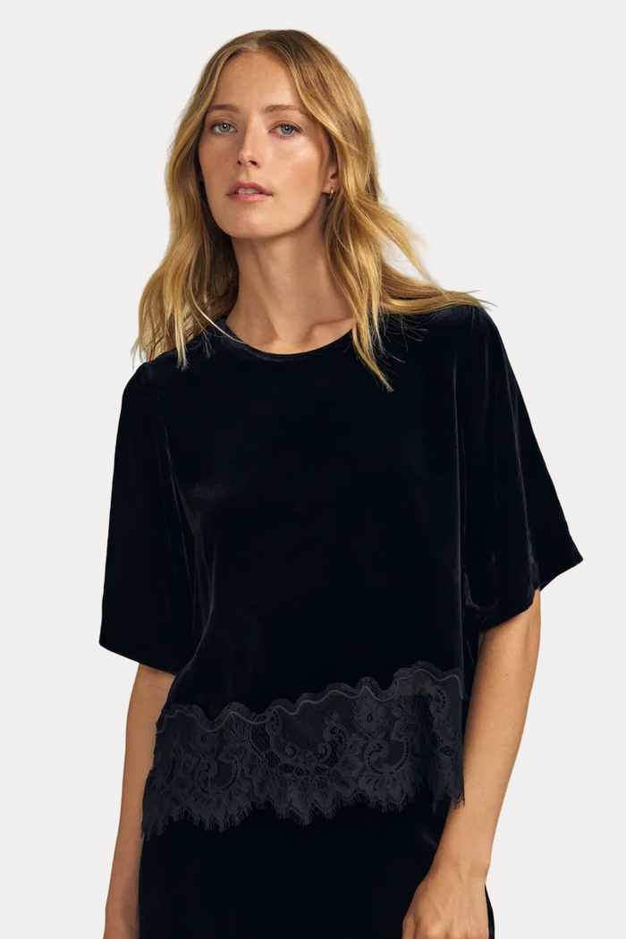 A woman modeling a black velvet short-sleeve top with a matching black lace waistband, set against a solid beige studio background.