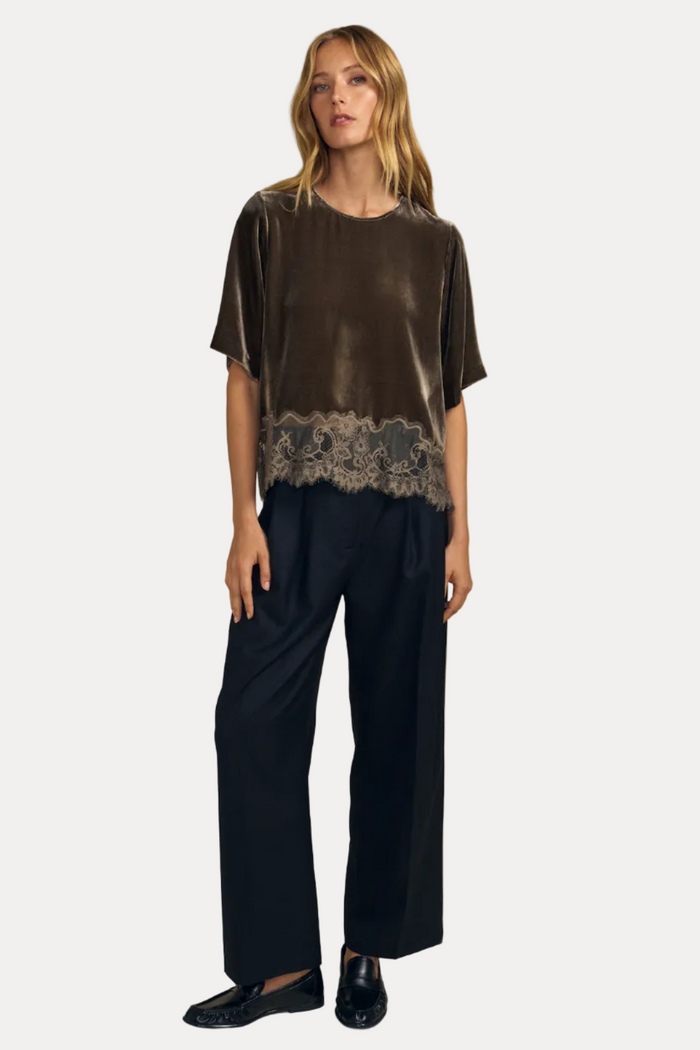 A woman modeling a short-sleeve top made of dark brown velvet with a lace hem, paired with dark wide-leg trousers and black loafers, against a solid beige background.