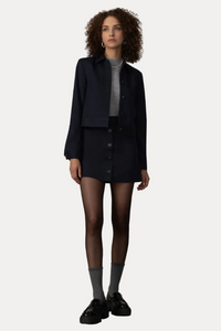 A model stands in front of a beige curtain backdrop wearing a dark navy cropped jacket and matching button-front mini skirt over a gray mock-neck top, paired with gray socks and black loafers.