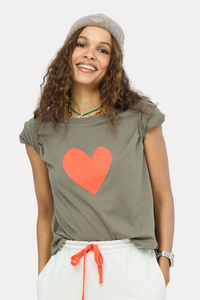 A woman wearing a cropped olive green t-shirt with a large orange heart on the front, paired with white sweatpants and a gray knit beanie, set against a light blue studio background.