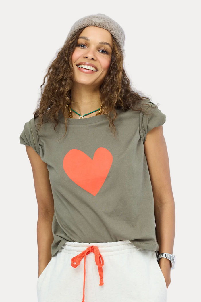 A woman wearing a cropped olive green t-shirt with a large orange heart on the front, paired with white sweatpants and a gray knit beanie, set against a light blue studio background.