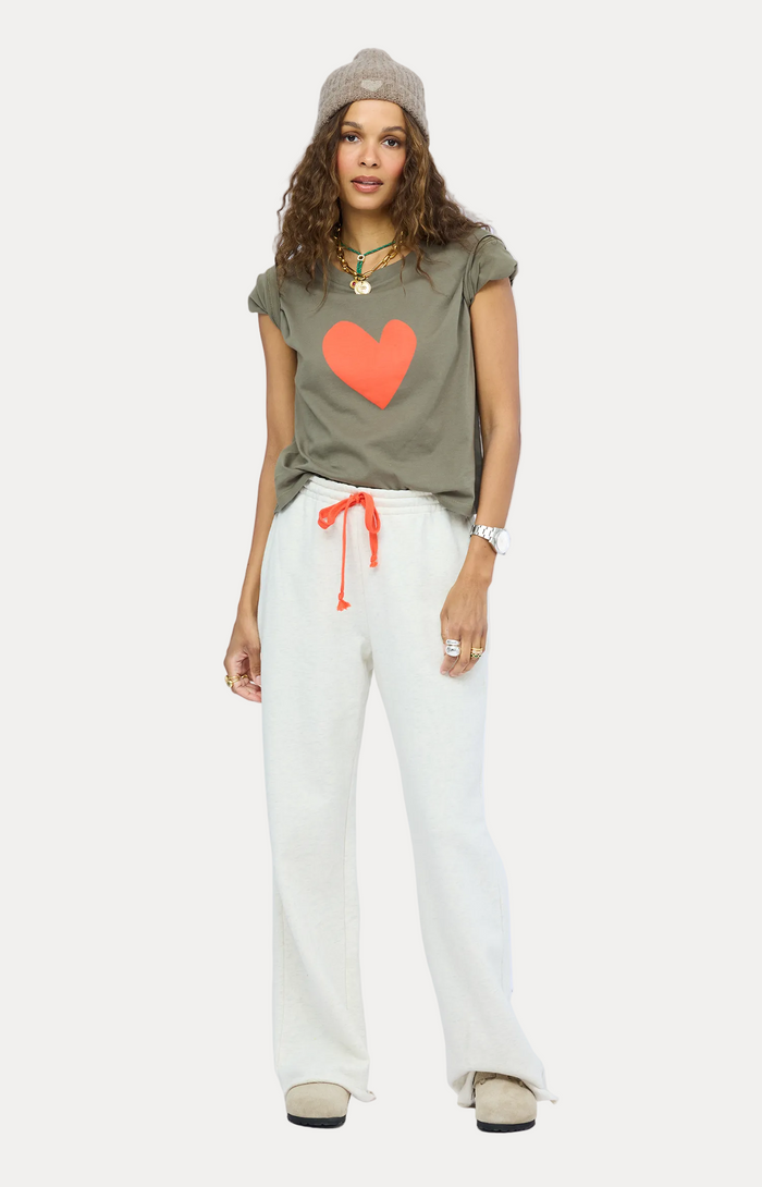 A woman modeling a casual outfit consisting of an olive green t-shirt with a large orange heart graphic, cream wide-leg sweatpants with an orange drawstring, beige shoes, and a gray knit beanie against a light gray studio background.