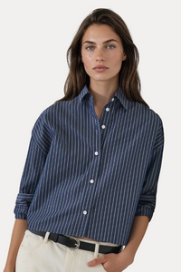 A woman wearing a navy blue button-down shirt with thin white vertical pinstripes, paired with off-white trousers and a black belt, standing against a plain off-white wall.