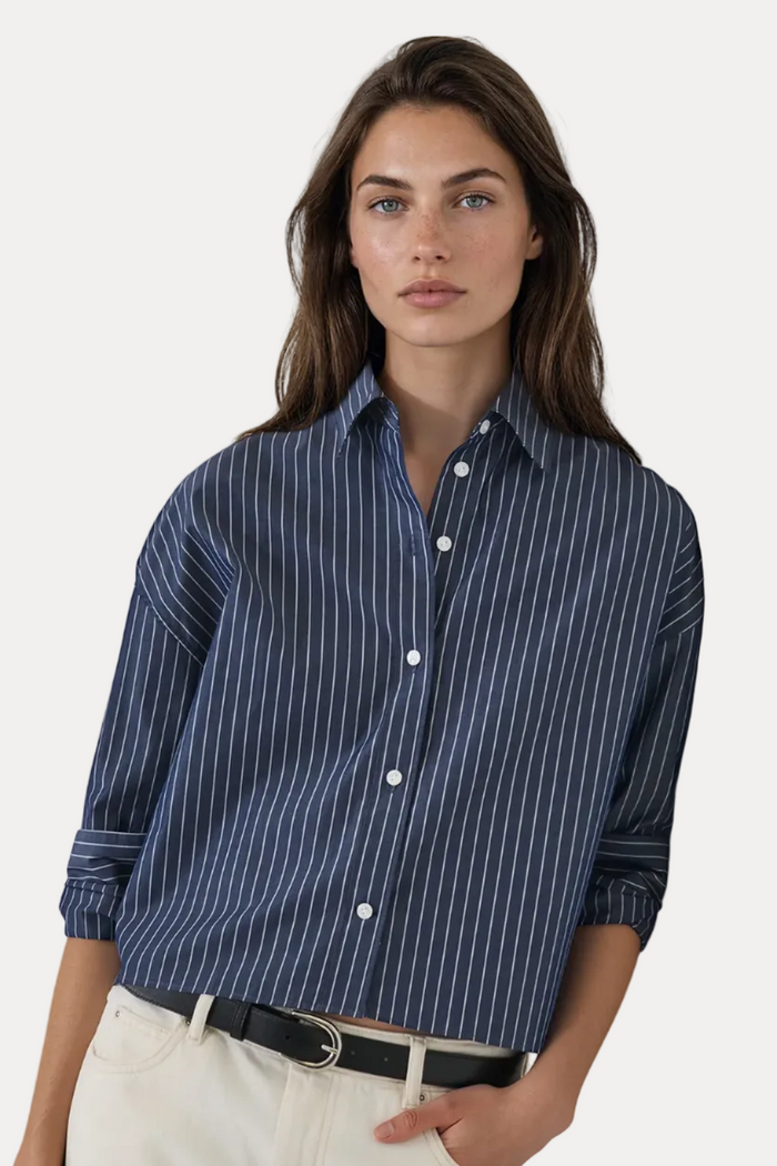 A woman wearing a navy blue button-down shirt with thin white vertical pinstripes, paired with off-white trousers and a black belt, standing against a plain off-white wall.