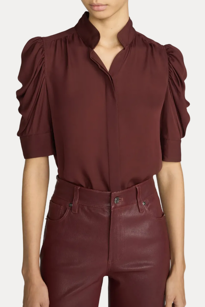 A model wearing a dark burgundy short-sleeve blouse with dramatic puff sleeves and a high collar, paired with matching faux-leather trousers, against a light gray studio background.