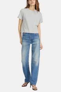 A model wearing a light gray short-sleeve crop top, blue wide-leg jeans with a faded wash, and black strappy high-heeled sandals, standing against a light gray studio background.
