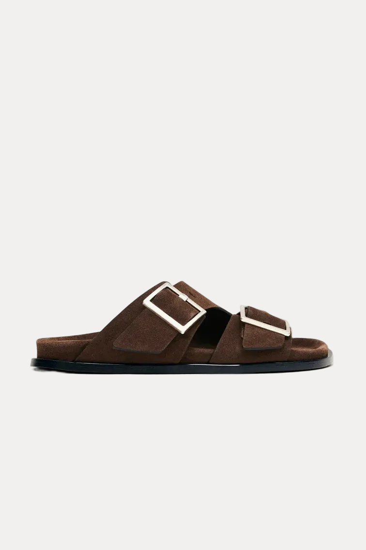 A pair of dark brown suede slide sandals with wide double straps and large silver-tone square buckles, shown in profile against a light off-white background.