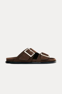 A pair of dark brown suede slide sandals with wide double straps and large silver-tone square buckles, shown in profile against a light off-white background.