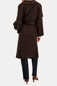 A woman shown from the back wearing a dark brown belted trench coat over dark blue jeans and brown heels, photographed against a plain white studio background.