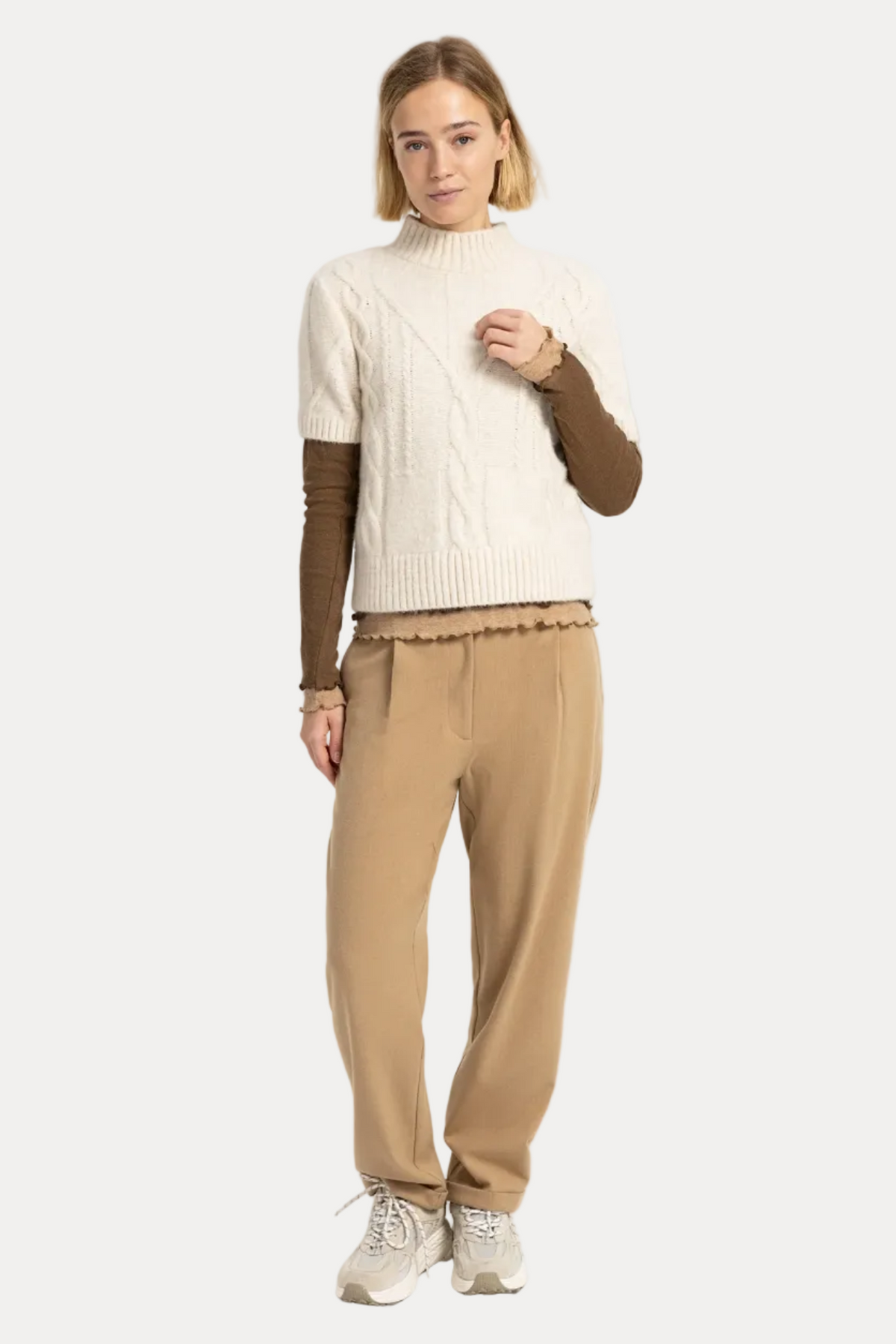 A woman modeling a cream cable-knit mock-neck sweater vest over a brown long-sleeve top, paired with beige trousers and white sneakers, against a plain white studio background.